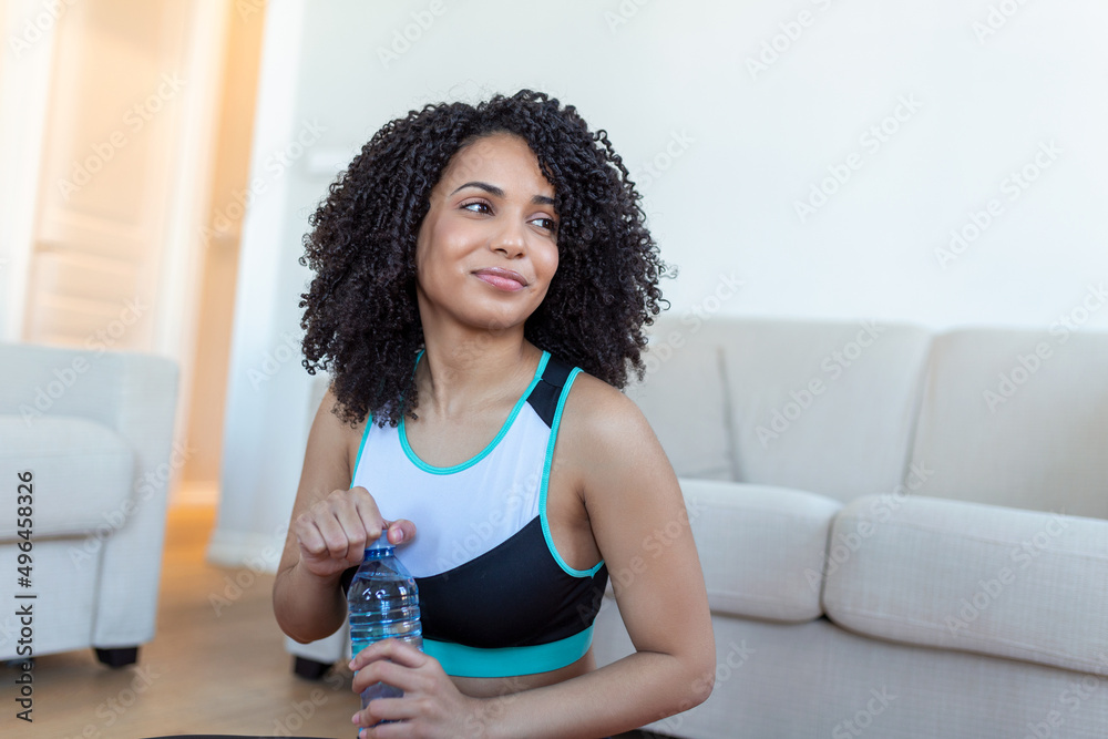 Young adult African American woman drinking water from plastic bottle, sitting on fitness mat and resting after training at home