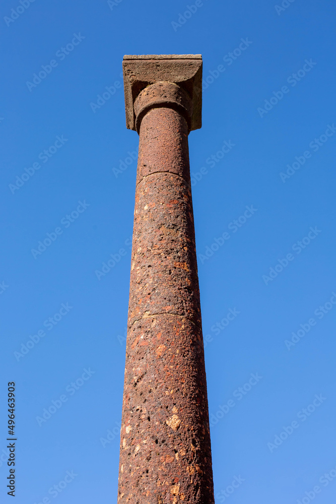 An ancient Roman column of volcanic rock is against a blue sky.
