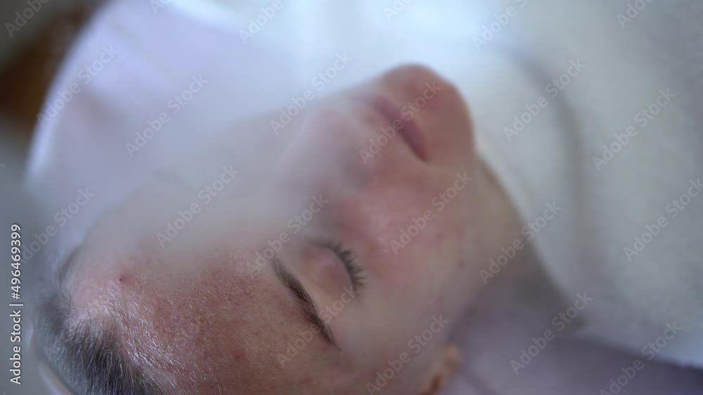 Close up of young woman with eyes closed having thermal steam treatment on problematic skin. Girl wearing white gown laying on couch in cosmetic salon Close up of young woman with eyes closed having thermal steam treatment on problematic skin. Girl wearing white gown laying on couch in cosmetic salon