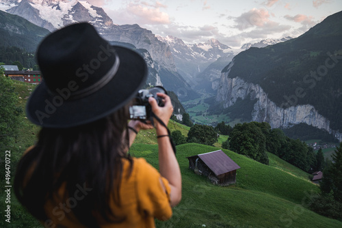 Young woman tourist taking photos with digital camera at famous touristic place in Wengen during sunset. Famous Instagram hiking place with green fields and snowy mountains over Lauterbrunnen Valley