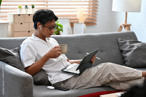 A portrait of an Asian man holding a cup of coffee and working on a tablet, for business, education, home and technology concept.