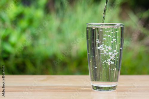 Pour water into glass on wooden table outdoors and green background. © Copteranansak