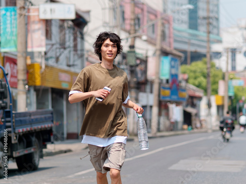 Portrait of handsome Chinese young man with black curly hair in brown T-shirt and pants running on Shanghai old town street with bottle of water in hand, front view of cool young man.
