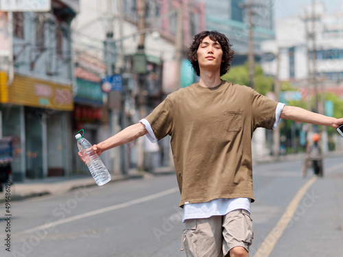 Portrait of handsome Chinese young man with black curly hair in brown T-shirt and pants walking on Shanghai old town street with arms up and eyes closed, water and mobile phone in hand, looks tired.
