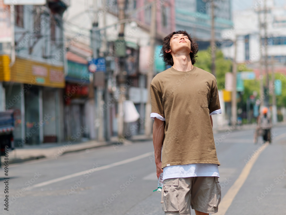 Portrait of handsome Chinese young man with black curly hair in brown T-shirt and pants walking on Shanghai old town street with head up and eyes closed, looks tired.