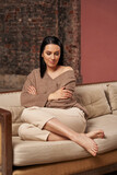 a beautiful woman with black hair in cozy clothes of a natural shade sits on the sofa with her legs crossed and hugging herself by the shoulders