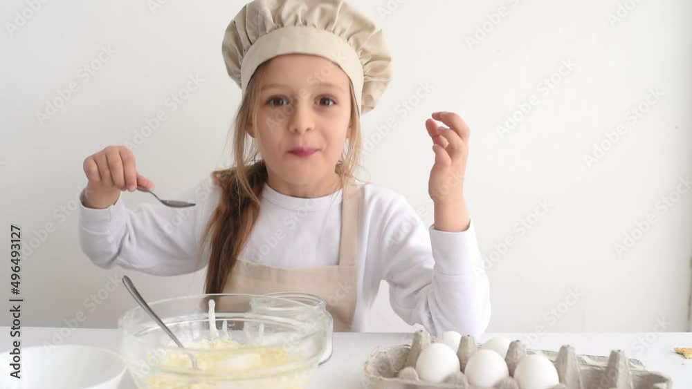 Small cute girl with smile eating sugar and enjoys. Kid prepares dough for cake. On side stand eggs and in bowl sugar on white table. Small cute girl with smile eating sugar and enjoys. Kid prepares dough for cake. On side stand eggs and in bowl sugar on white table.