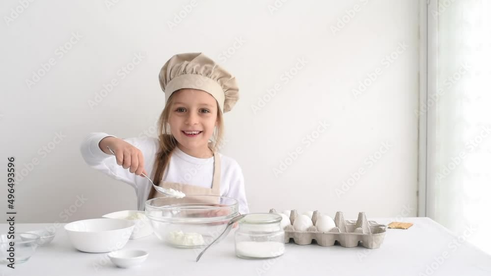Small cute girl with amazing smile prepares dough for cake. Kid in chef's hat puts white cream in bowl. Child enjoy and learning to cook. On white table stand eggs and in bowls sugar, salt and soda Small cute girl with amazing smile prepares dough for cake. Kid in chef's hat puts white cream in bowl. Child enjoy and learning to cook. On white table stand eggs and in bowls sugar, salt and soda