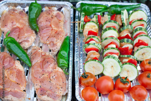 Slices of raw pork neck, hot green chilli pepper and chicken and vegetable skewers lying on an aluminum tray and waiting to be grilled.
