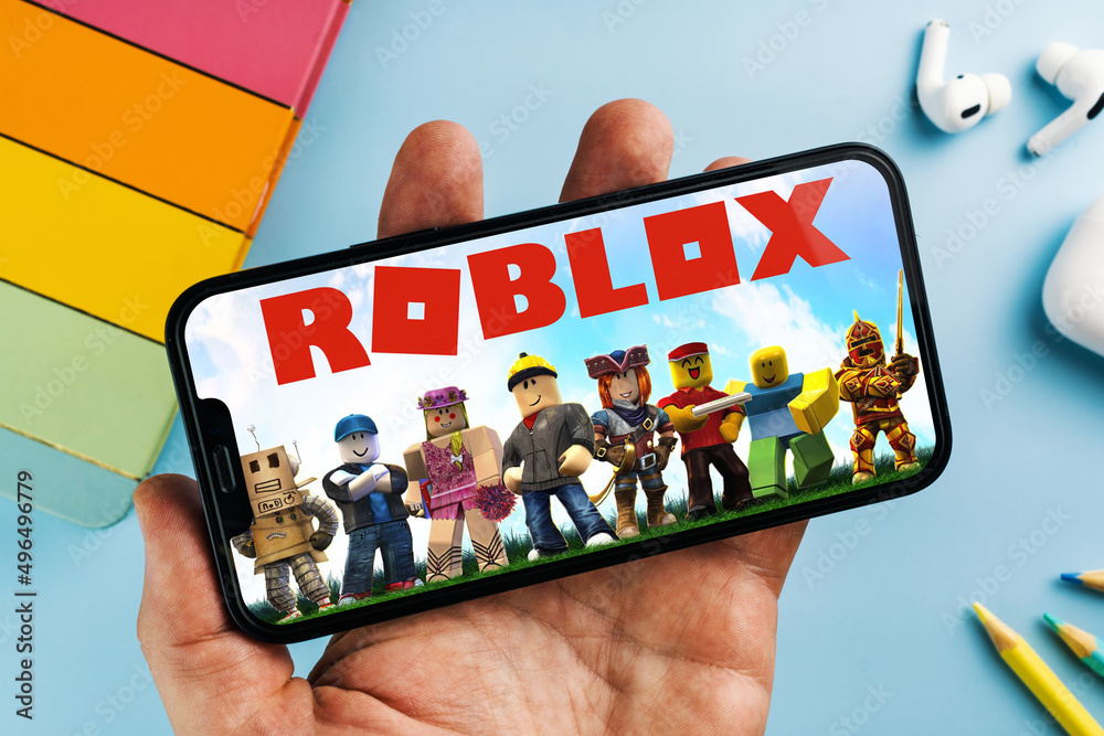 Man holding a smartphone with Roblox mobile game app on the screen. Blue  background with school supplies, AirPods, video game controller. Rio de  Janeiro, RJ, Brazil. April 2022 Stock Photo