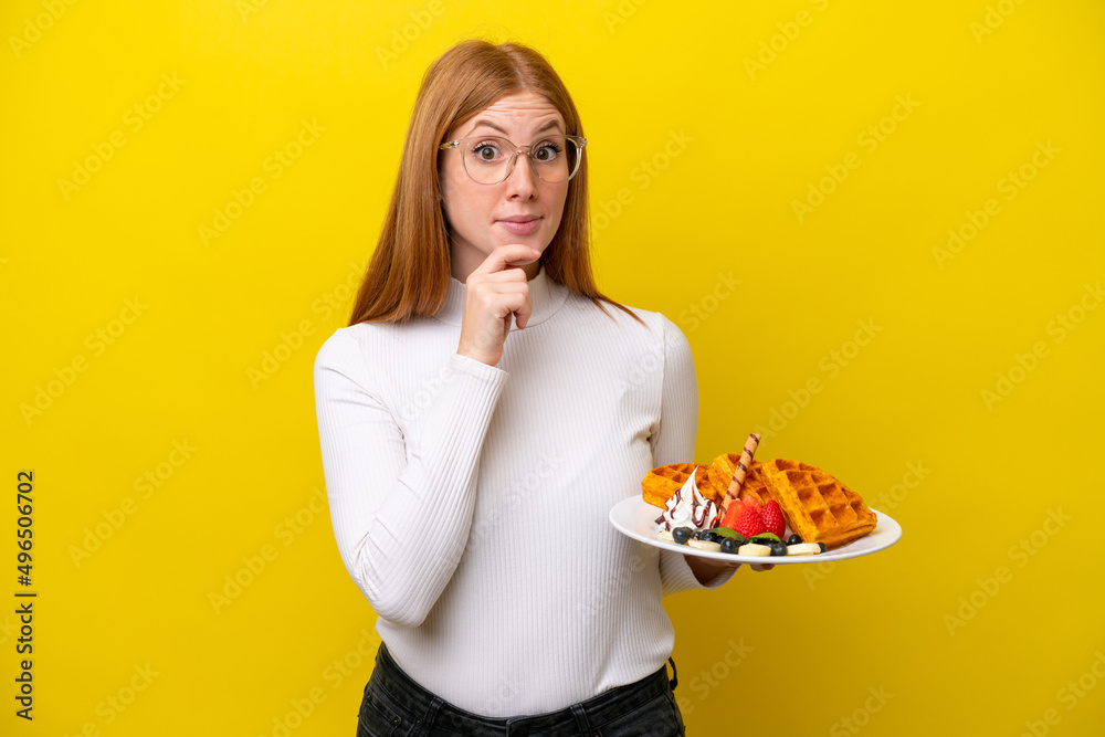 Young redhead woman holding waffles isolated on yellow background having doubts and thinking Young redhead woman holding waffles isolated on yellow background having doubts and thinking