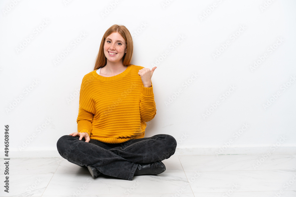 Young redhead woman sitting on the floor isolated on white background pointing to the side to present a product