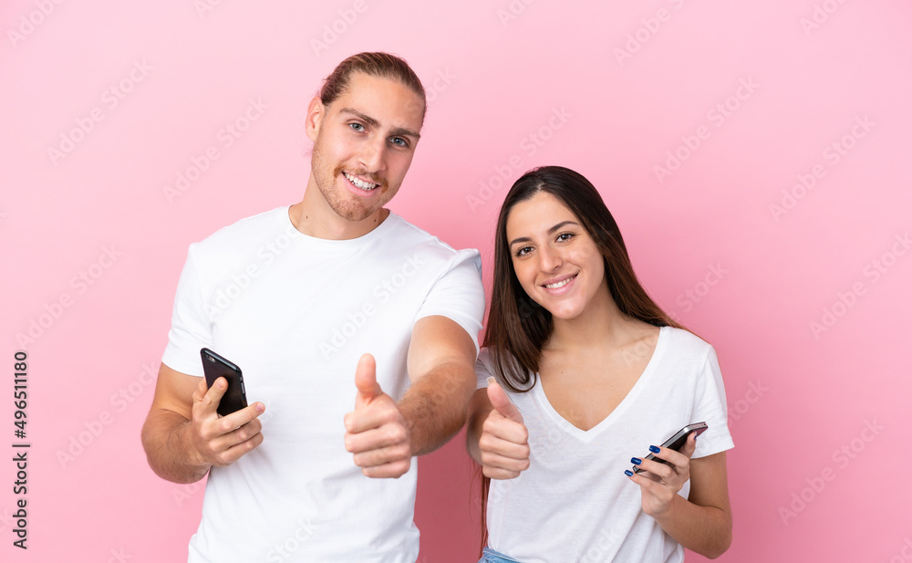 Young caucasian couple isolated on pink background using mobile phone while doing thumbs up Young caucasian couple isolated on pink background using mobile phone while doing thumbs up