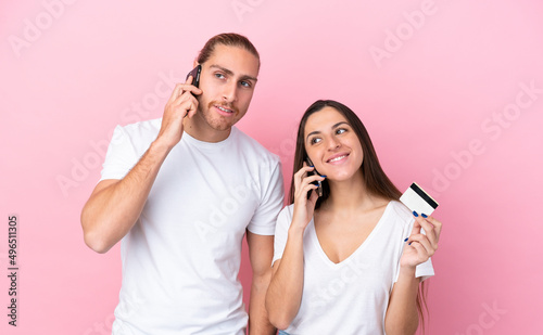 Young caucasian couple isolated on pink background keeping a conversation with the mobile phone and holding a credit card