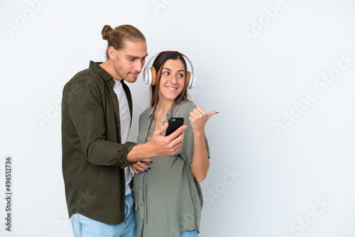 Young caucasian couple isolated on white background pregnant and listening music while pointing side