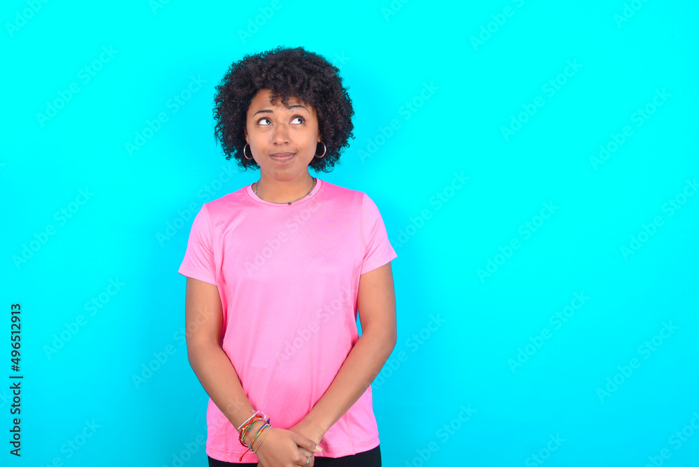 young girl with afro hairstyle wearing pink T-shirt over blue background looking aside into empty space thoughtful young girl with afro hairstyle wearing pink T-shirt over blue background looking aside into empty space thoughtful