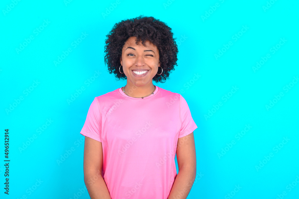 Coquettish young girl with afro hairstyle wearing pink T-shirt over blue background smiling happily, blinking at camera in a playful manner, flirting with you. Coquettish young girl with afro hairstyle wearing pink T-shirt over blue background smiling happily, blinking at camera in a playful manner, flirting with you.