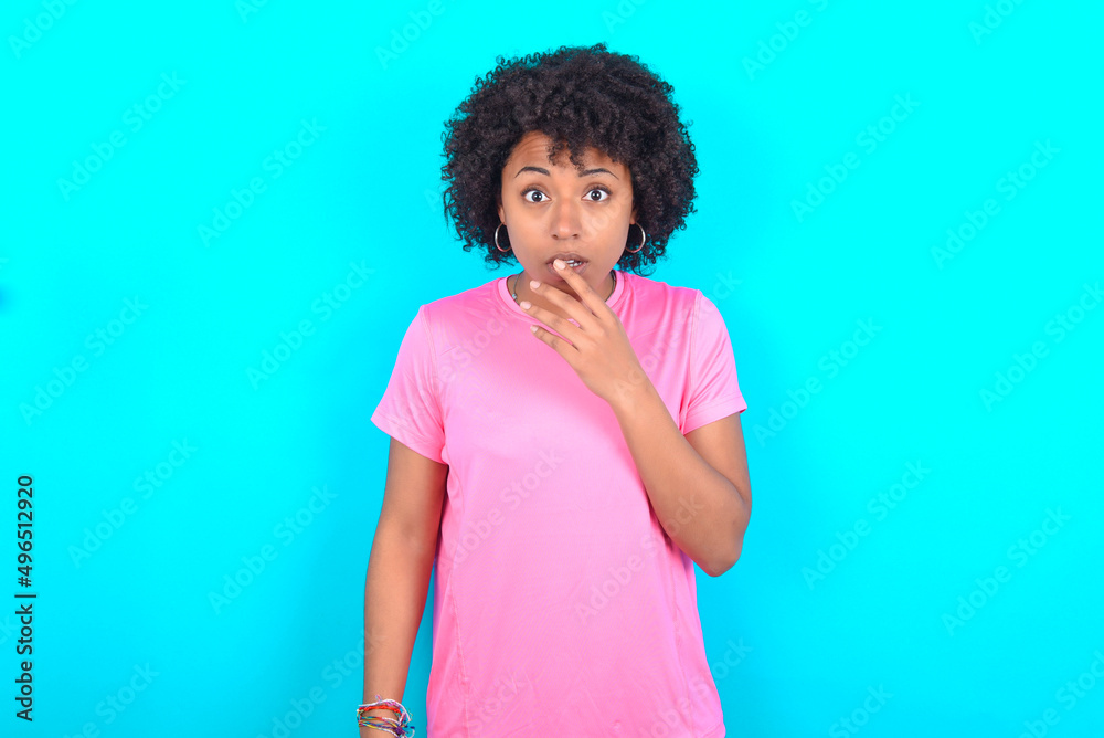 Nervous puzzled young girl with afro hairstyle wearing pink T-shirt over blue background opens mouth from surprise, reacts on sudden news.