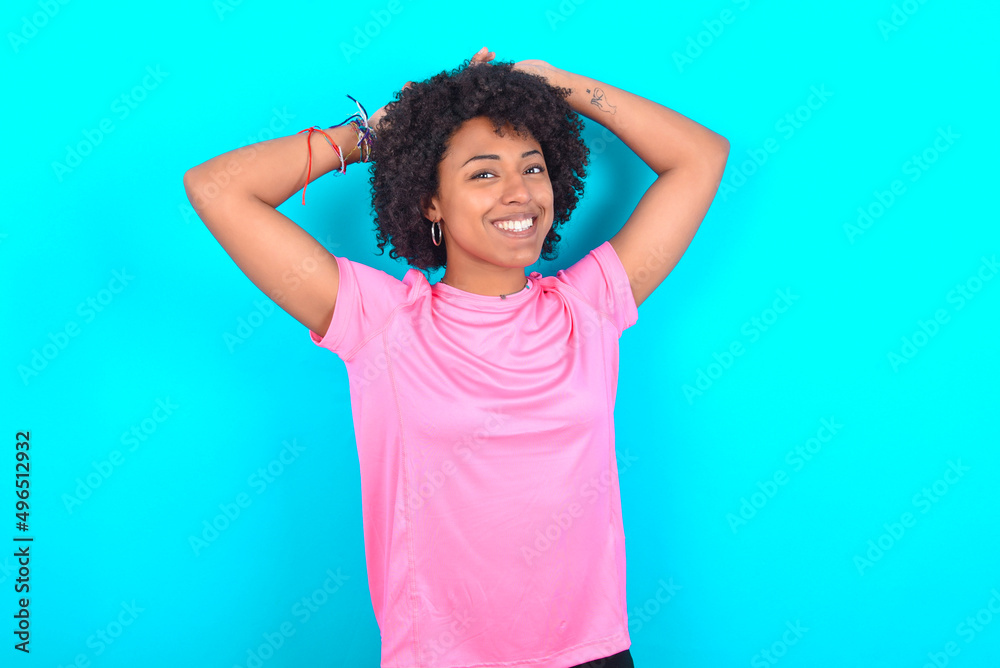 young girl with afro hairstyle wearing pink T-shirt over blue background stretching arms, relaxed position.