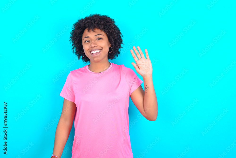 young girl with afro hairstyle wearing pink T-shirt over blue background Waiving saying hello happy and smiling, friendly welcome gesture.