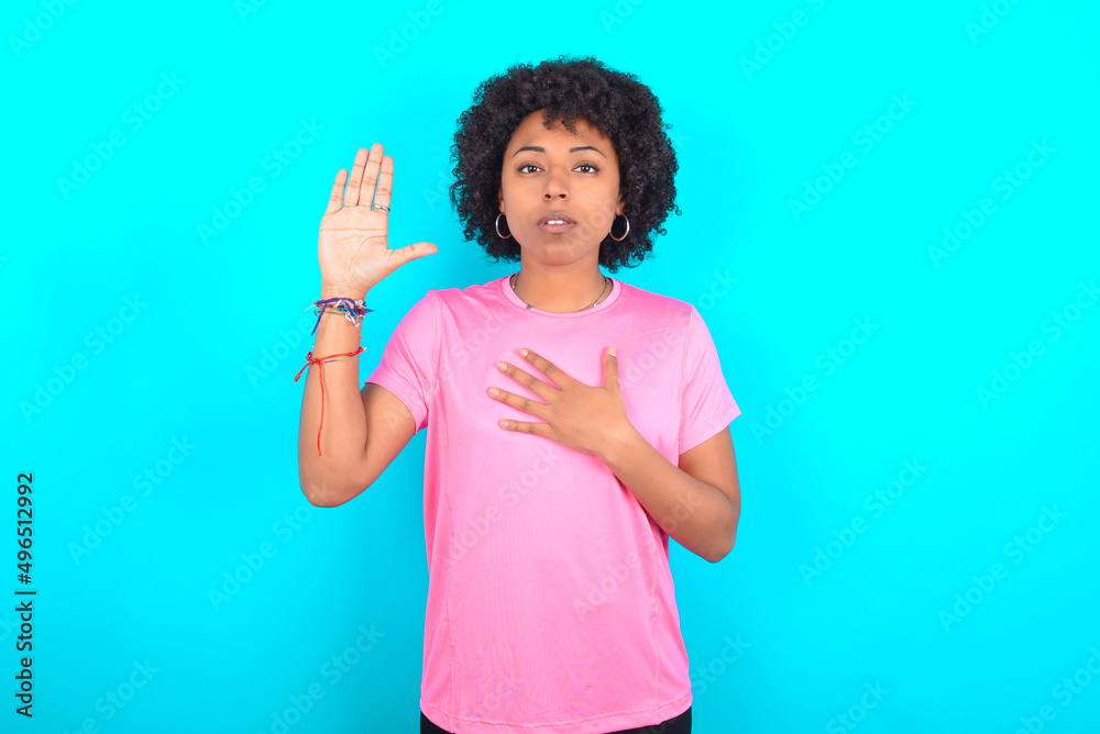 young girl with afro hairstyle wearing pink T-shirt over blue background Swearing with hand on chest and open palm, making a loyalty promise oath young girl with afro hairstyle wearing pink T-shirt over blue background Swearing with hand on chest and open palm, making a loyalty promise oath