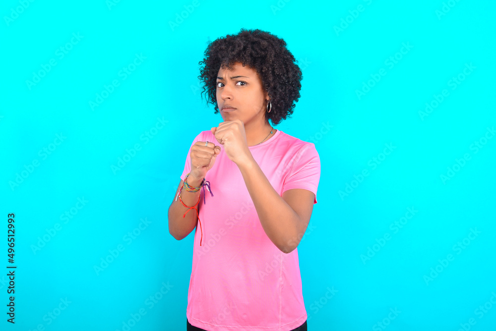 young girl with afro hairstyle wearing pink T-shirt over blue background Ready to fight with fist defense gesture, angry and upset face, afraid of problem. young girl with afro hairstyle wearing pink T-shirt over blue background Ready to fight with fist defense gesture, angry and upset face, afraid of problem.