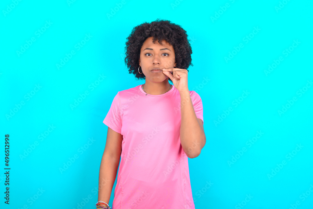 young girl with afro hairstyle wearing pink T-shirt over blue background mouth and lips shut as zip with fingers. Secret and silent, taboo talking. young girl with afro hairstyle wearing pink T-shirt over blue background mouth and lips shut as zip with fingers. Secret and silent, taboo talking.