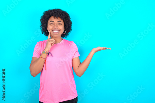 Funny young girl with afro hairstyle wearing pink T-shirt over blue background hold open palm new product great proposition photo