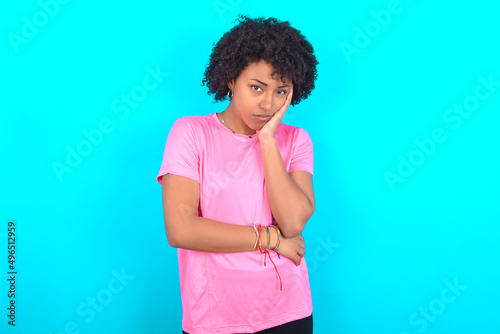 Very bored young girl with afro hairstyle wearing pink T-shirt over blue background holding hand on cheek while support it with another crossed hand, looking tired and sick,