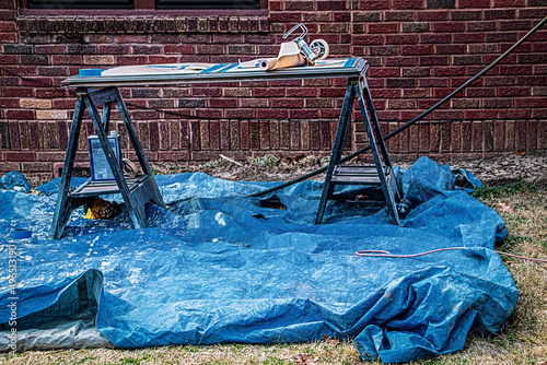 Plastic sawhorse table set up outside brick house on blue tarp with painting supplies on it and electric cords for some project