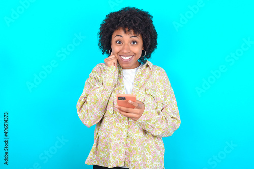 Joyous young girl with afro hairstyle wearing floral shirt over blue background poses with mobile phone device, types text message on modern smartphone, watches funny video during free time, 