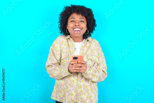 Excited young girl with afro hairstyle wearing floral shirt over blue background holding smartphone and looking amazed to the camera after receiving good news. photo