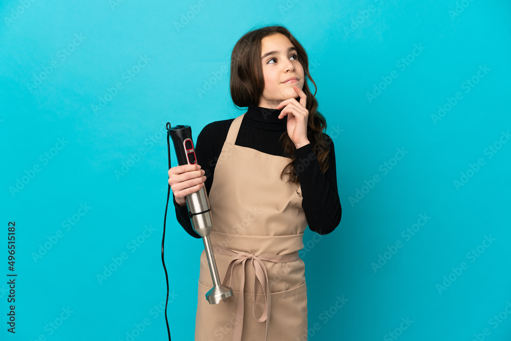 Little girl using hand blender isolated on blue background having doubts while looking up Little girl using hand blender isolated on blue background having doubts while looking up