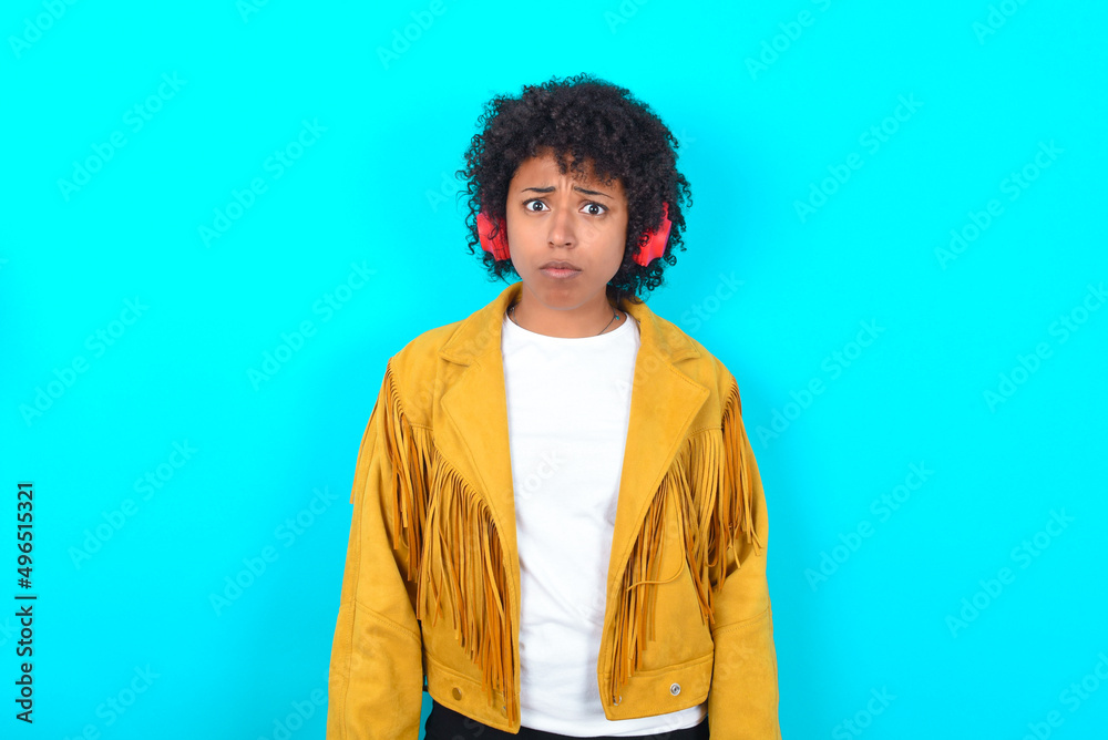 Serious displeased Young woman with afro hairstyle wearing yellow fringe jacket over blue background looks puzzled at camera being angry wears stereo headphones listens music while walking at street Serious displeased Young woman with afro hairstyle wearing yellow fringe jacket over blue background looks puzzled at camera being angry wears stereo headphones listens music while walking at street