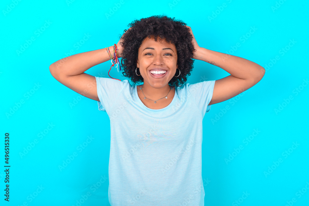 Cheerful overjoyed Young woman with afro hairstyle wearing blue T-shirt over blue background reacts rising hands over head after receiving great news.