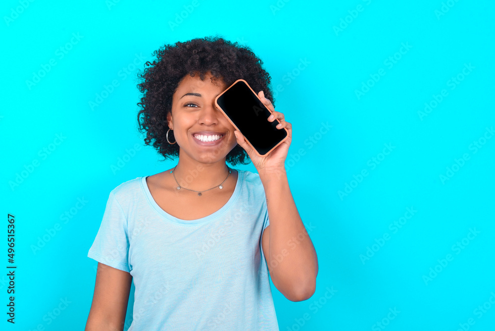 Young woman with afro hairstyle wearing blue T-shirt over blue background holding modern smartphone covering one eye while smiling Young woman with afro hairstyle wearing blue T-shirt over blue background holding modern smartphone covering one eye while smiling