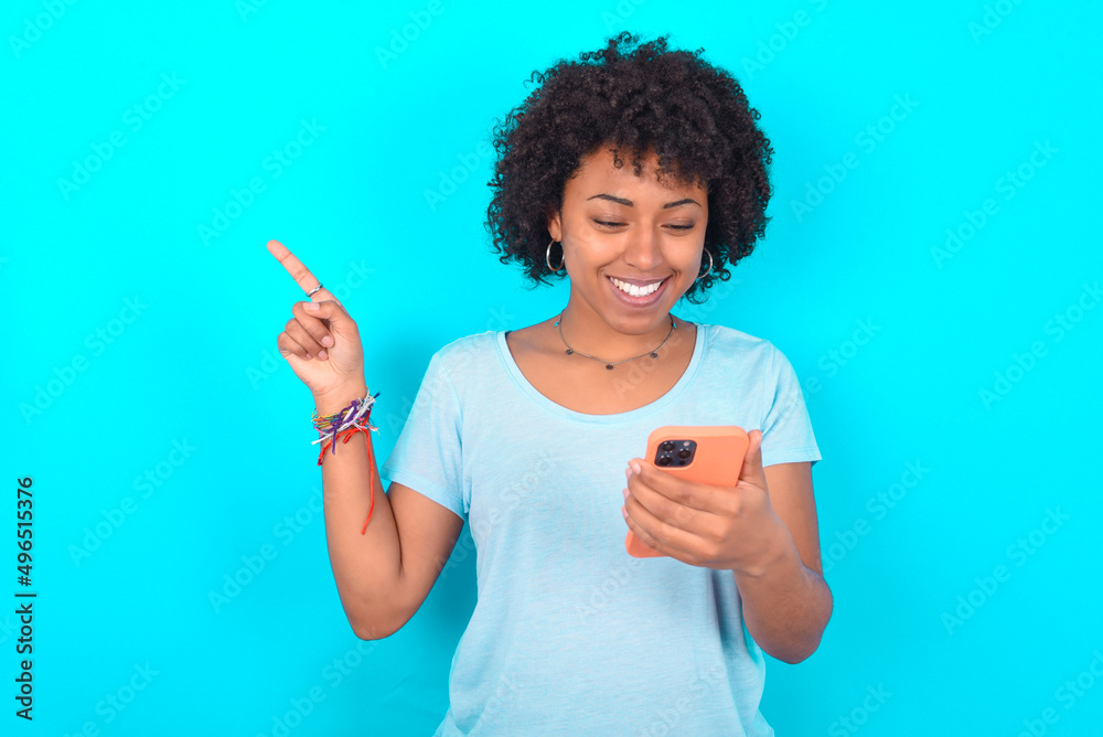 Smiling Young woman with afro hairstyle wearing blue T-shirt over blue background pointing finger at blank space holding phone in one hand Smiling Young woman with afro hairstyle wearing blue T-shirt over blue background pointing finger at blank space holding phone in one hand