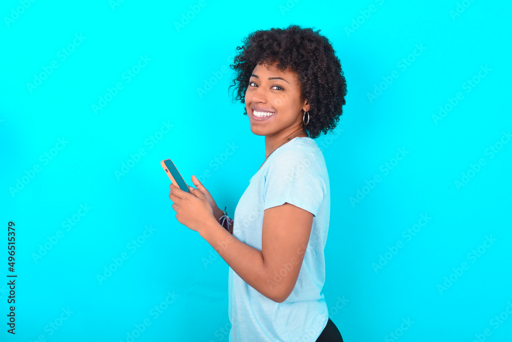 Rear view photo portrait of Young woman with afro hairstyle wearing blue T-shirt over blue background using smartphone smiling Rear view photo portrait of Young woman with afro hairstyle wearing blue T-shirt over blue background using smartphone smiling