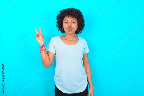 Young woman with afro hairstyle wearing blue T-shirt over blue background makes peace gesture keeps lips folded shows v sign. Body language concept photo