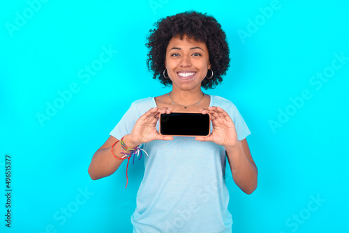 Cheerful cheery content Young woman with afro hairstyle wearing blue T-shirt over blue background holding in hands device hobby smm post blog