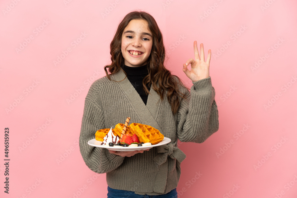 Little girl holding waffles isolated on pink background showing ok sign with fingers Little girl holding waffles isolated on pink background showing ok sign with fingers