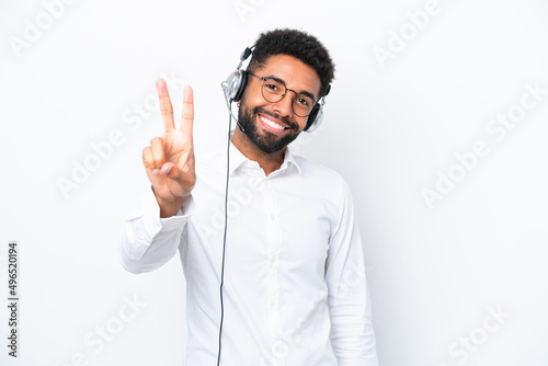 Telemarketer Brazilian man working with a headset isolated on white background smiling and showing victory sign
