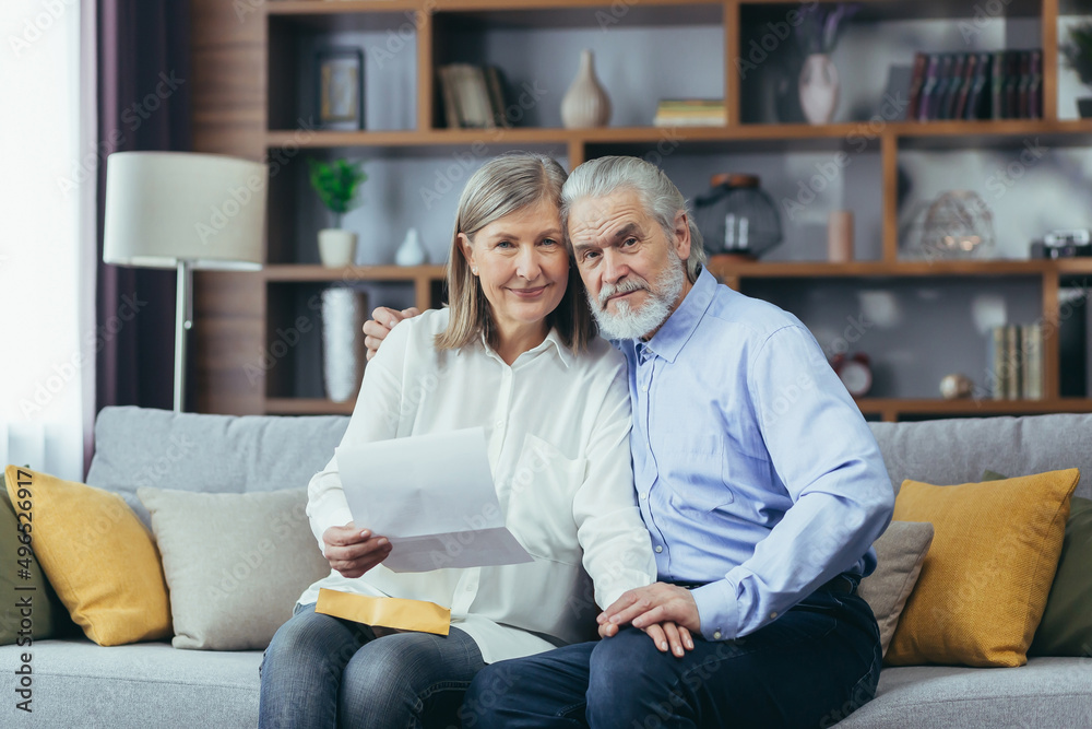 Senior couple man and woman together at home, received good news letter, happy and smiling sitting on sofa, looking at camera
