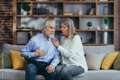 Senior mature woman helping her husband with chest pain. Old man feeling bad pain disease. Wife supporting husband - Elderly couple suffering from symptoms heart attack ache. sitting at home indoors