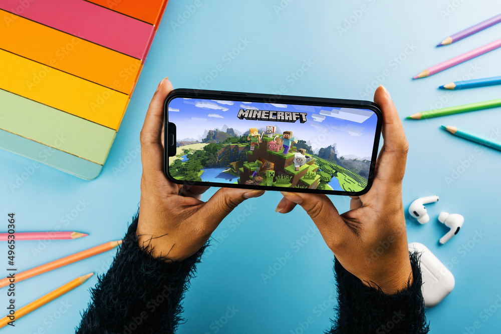 Fototapeta premium Girl holding a smartphone with Minecraft mobile game app on the screen. Blue background with school supplies, AirPods, video game controller. Rio de Janeiro, RJ, Brazil. April 2022