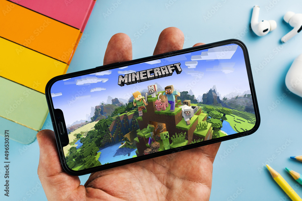 Fototapeta premium Man holding a smartphone with Minecraft mobile game app on the screen. Blue background with school supplies, AirPods, video game controller. Rio de Janeiro, RJ, Brazil. April 2022