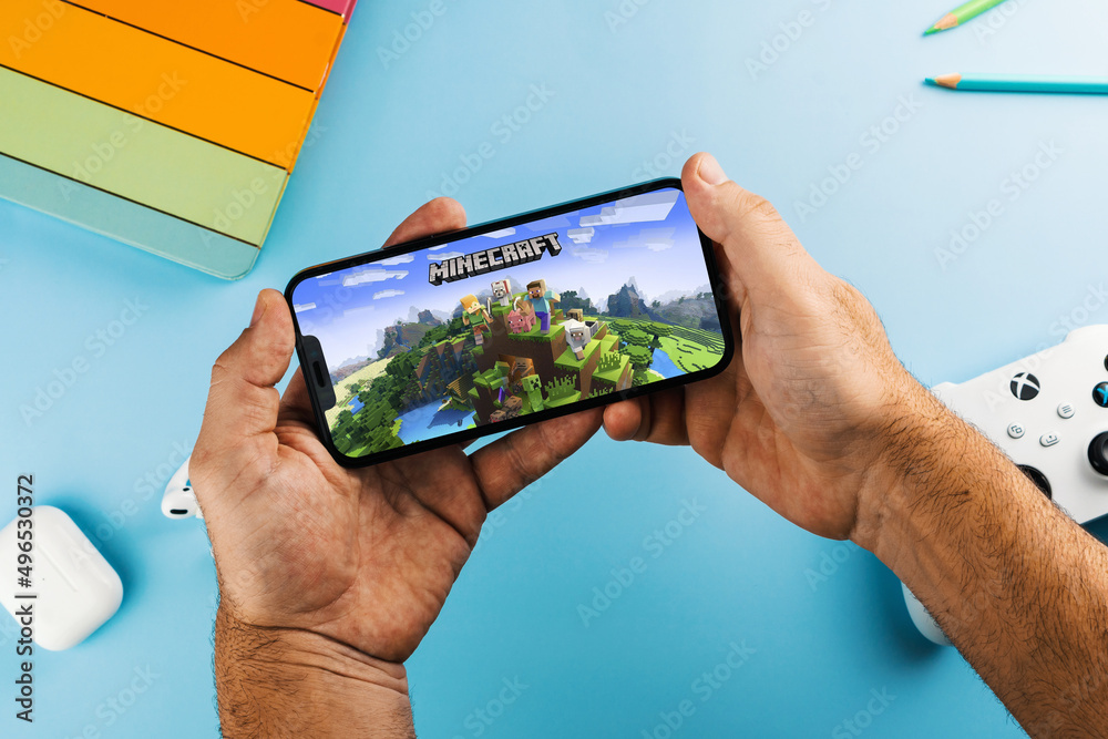 Fototapeta premium Man holding a smartphone with Minecraft mobile game app on the screen. Blue background with school supplies, AirPods, video game controller. Rio de Janeiro, RJ, Brazil. April 2022