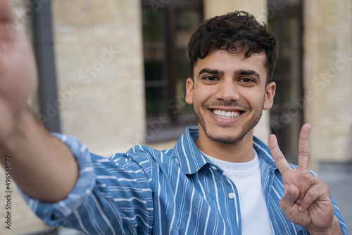 Handsome  smiling Indian man holding mobile phone taking selfie showing victory sign on the street. Happy stylish asian influencer communication online with subscribers outdoors