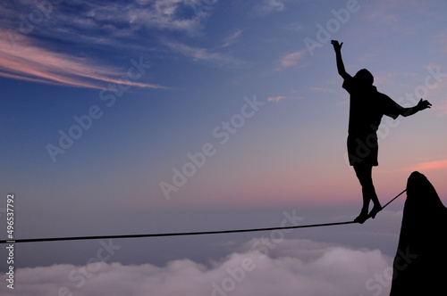 Silhouette of young man balancing on slackline high above clouds and mountains. Slackliner balancing on tightrope during sunset, highline silhouette.