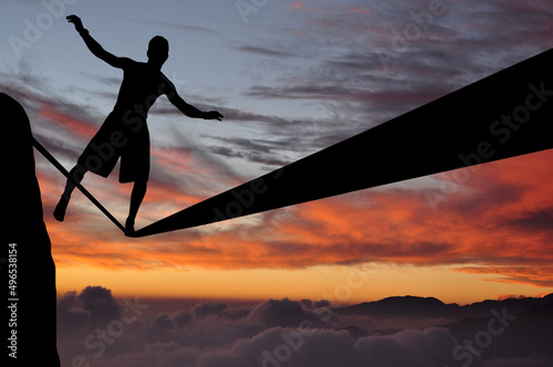 Silhouette of young man balancing on slackline high above clouds and mountains. Slackliner balancing on tightrope during sunset, highline silhouette.